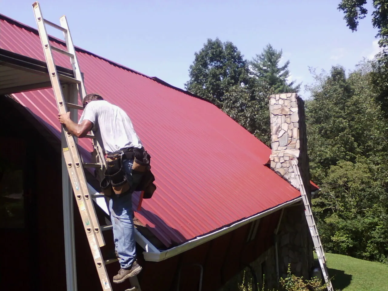 Roofer on a ladder beside a red metal roof and stone chimney — Soffit & Fascia Repair by Roofing Brothers in Barnstable
